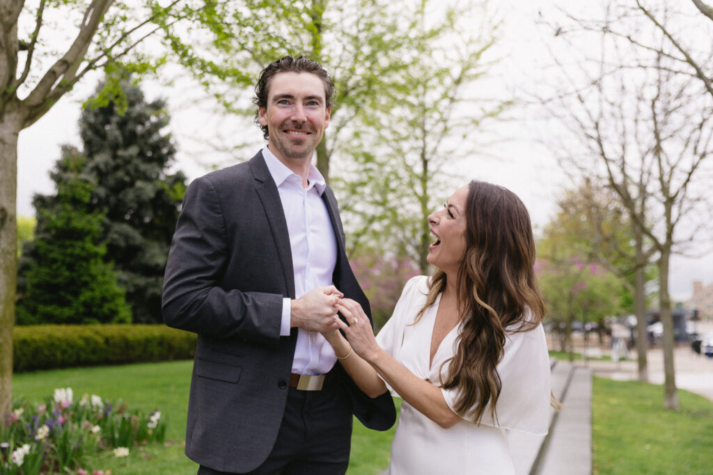 A man in a suit stands smiling while a woman in a white dress holds his hand and looks up at him, laughing, in a park with green trees and blooming flowers.