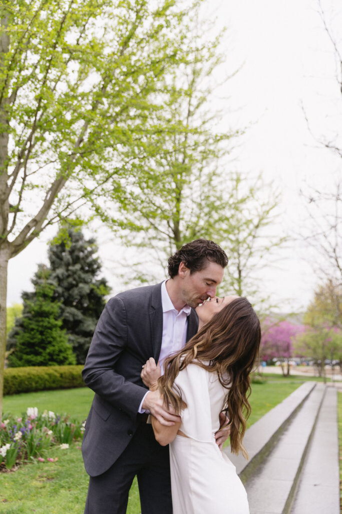 A man in a suit and a woman in a white dress share a romantic kiss outdoors, surrounded by green trees and blooming flowers along a garden path.