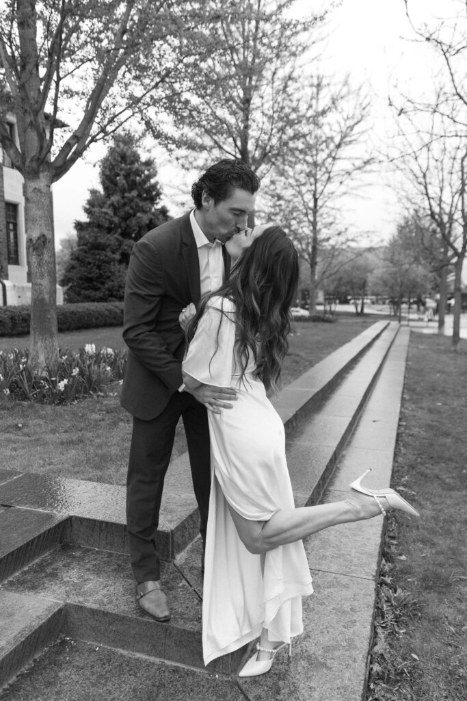 A man in a suit kisses a woman in a white dress as she lifts one leg, standing on stone steps outdoors amid trees and grass. The scene is romantic and candid, captured in black and white.