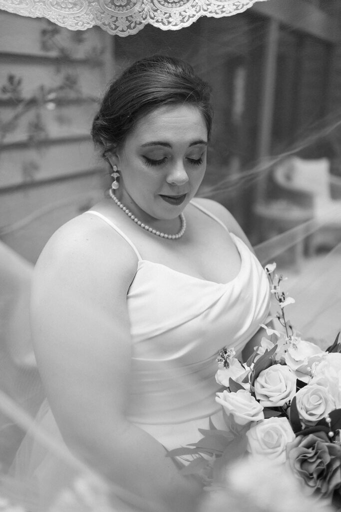 A bride in a white dress with a pearl necklace and earrings looks down, holding a bouquet of roses. The photo is black and white, with delicate lace visible at the top edge.
