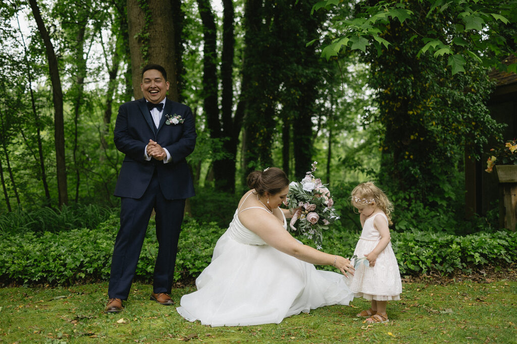 A bride in a white gown crouches and smiles at a young girl in a white dress, while a groom in a navy suit laughs nearby. They are outdoors in a green, wooded area.