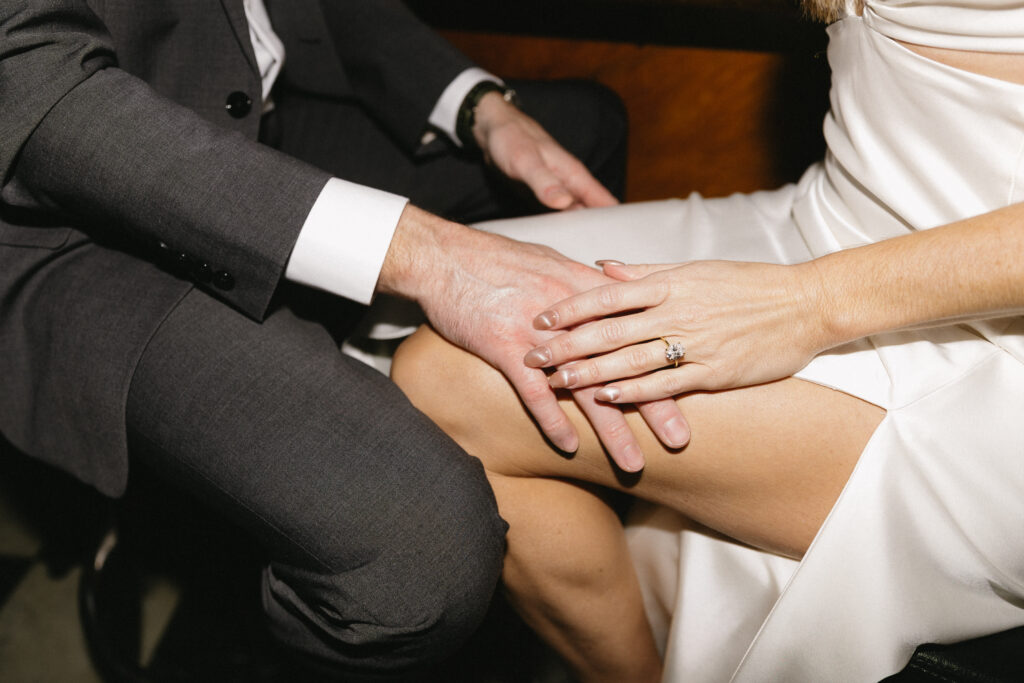 A close-up of a couple sitting together, with a man in a dark suit gently placing his hand on a womans knee. The woman is wearing a white dress and an engagement ring.