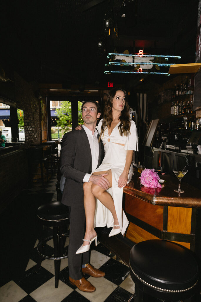 A woman in a white dress sits on a bar counter with a man in a suit standing beside her, his hand on her leg. They both look off into the distance in a dimly lit bar with checkered floors and a bouquet of pink flowers nearby.