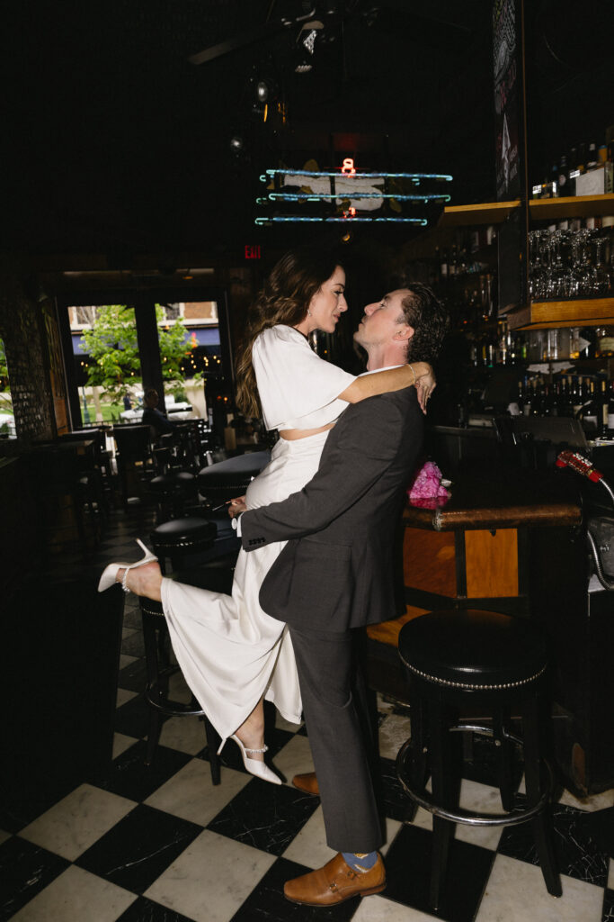 A man in a suit lifts a woman in a white dress off the ground as they smile at each other in a dimly lit bar with checkered floors and bottles lining the shelves.