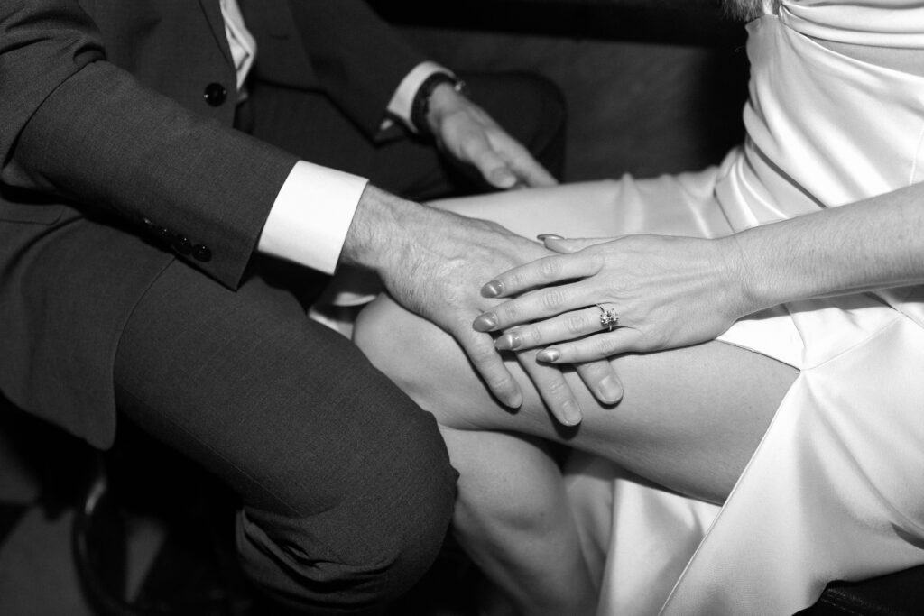 A black and white photo shows a man in a suit and a woman in a satin dress sitting close together, with their hands gently resting on her knee. The woman wears a ring on her left hand.