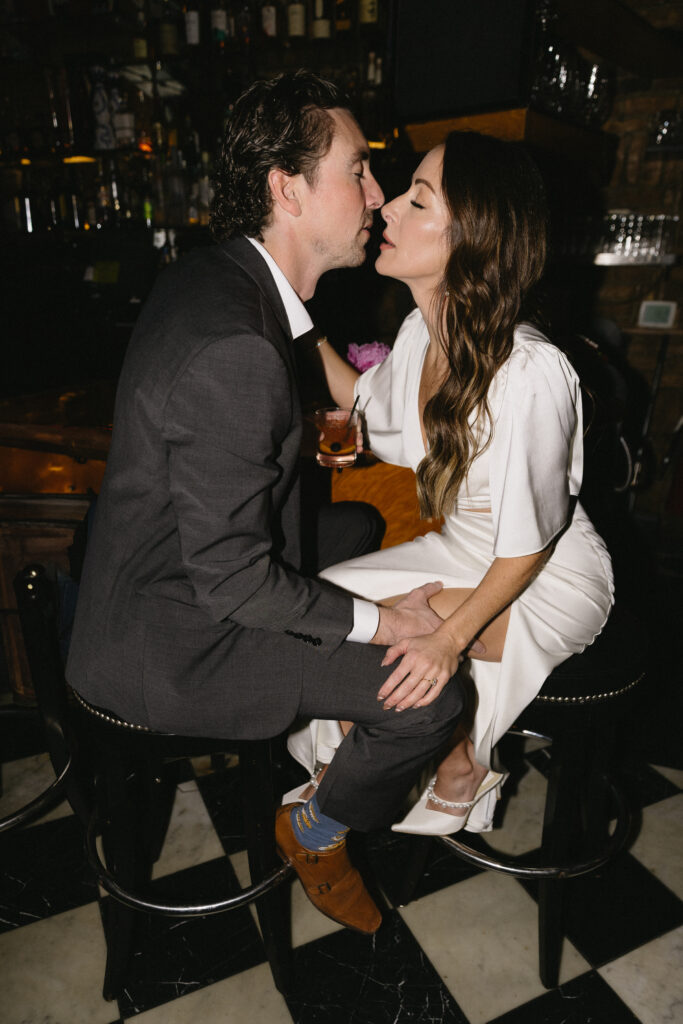 A man in a suit and a woman in a white dress sit closely on bar stools, holding hands and leaning in for a kiss at a dimly lit bar, with drinks in hand and checkered floor visible beneath them.