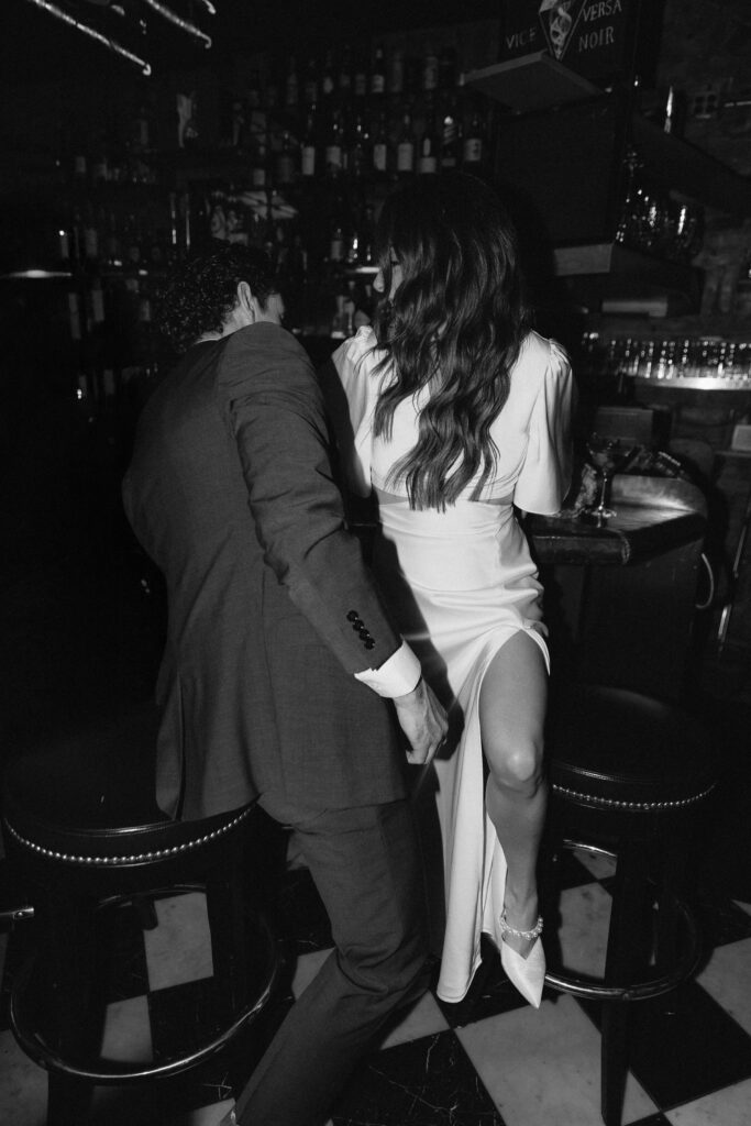 A man in a suit and a woman in a long white dress sit closely together on bar stools in a dimly lit bar. The woman’s dress has a high slit, and the two appear to be laughing and enjoying themselves.