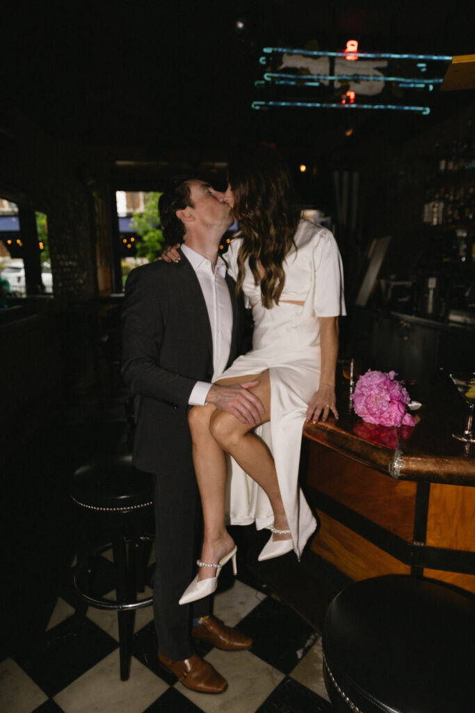 A couple dressed formally kiss at a dimly lit bar; the woman in a white dress sits on the counter with a bouquet of pink flowers beside her, while the man stands close, holding her waist.