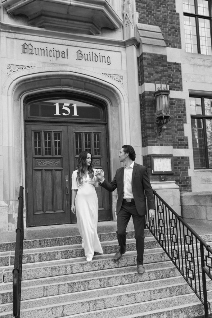 A couple dressed formally descends the steps of a municipal building, holding hands and smiling at each other. The buildings entrance is labeled Municipal Building with the number 151 above the doors.