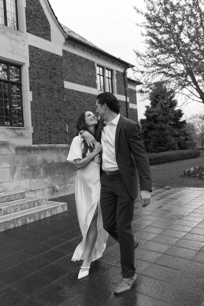 A couple dressed formally walk outside a brick building, smiling and looking at each other. The woman wears a light dress and the man wears a dark suit. Trees and steps are visible in the background.