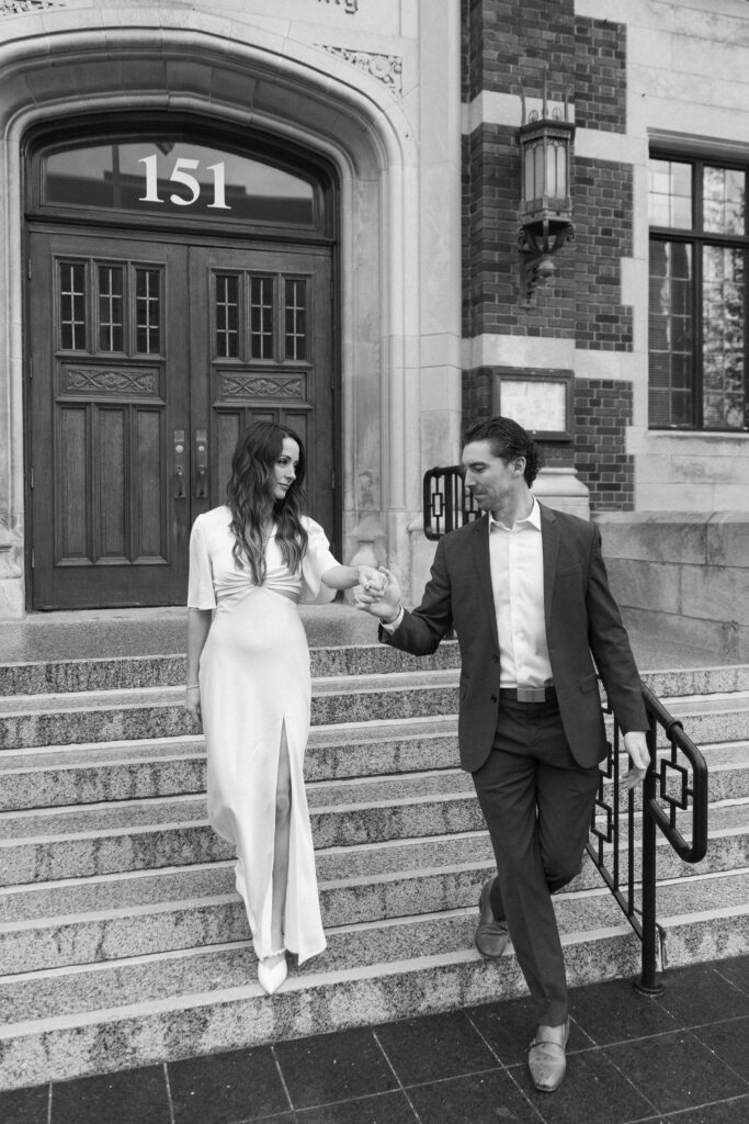 A woman in a white dress walks down stairs while holding hands with a man in a suit. They are in front of an ornate building with the number 151 above the door. The scene is in black and white.