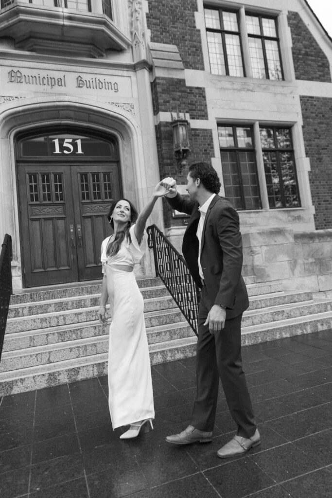 A couple dressed formally dances outside a building labeled Municipal Building with the address 151. The woman wears a long white dress, and the man is in a suit, holding her hand as she twirls. The image is in black and white.