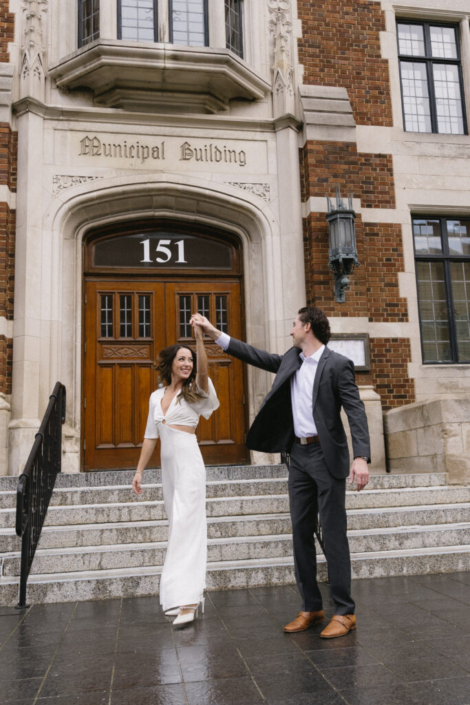 A man in a suit twirls a woman in a white dress on the steps of a historic municipal building with arched wooden doors and a stone facade.