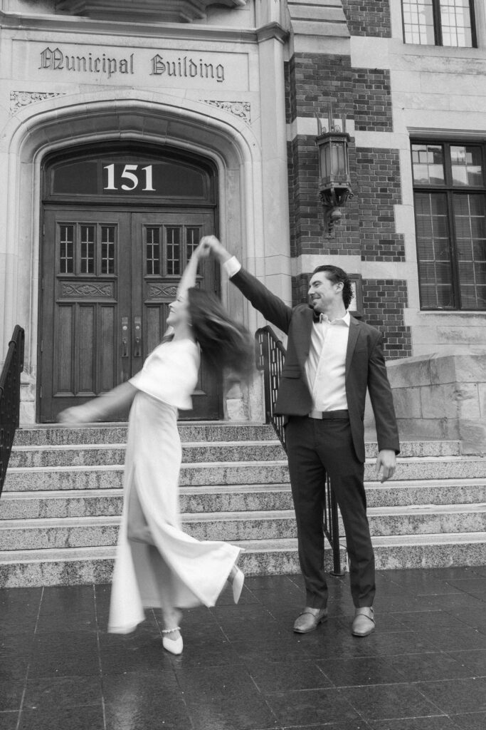 A man in a suit twirls a woman in a flowing dress outside a building labeled Municipal Building 151. The woman is mid-spin, her hair and dress flying, while both appear joyful.