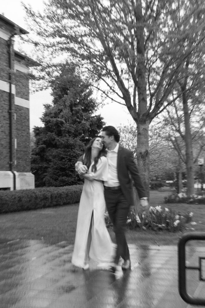 A couple dressed formally walks arm in arm on a wet sidewalk, smiling and looking at each other, with trees and a brick building in the background. The photo is black and white and has a romantic, candid feel.