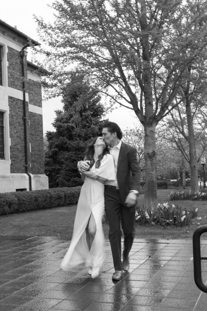 A couple dressed formally walk outside on a rainy day, laughing and embracing affectionately. Tall trees and a brick building are visible in the background. The scene is lively and romantic. Black and white photo.