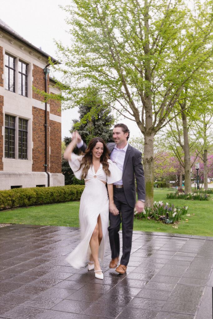 A couple dressed in formal attire dances joyfully on a wet sidewalk outside a brick building, surrounded by green trees and blooming flowers on a cloudy day.