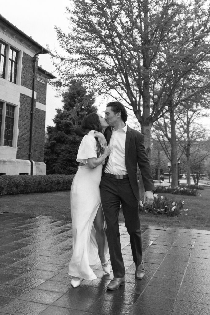 A couple dressed formally shares a kiss while walking outside on a wet sidewalk. The woman wears a white dress and heels; the man wears a suit. Trees and a large brick building are visible in the background.