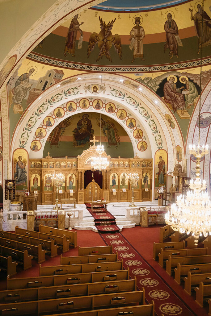 An ornate Orthodox church interior with rows of wooden pews, a red carpet, gold chandeliers, and an elaborate iconostasis decorated with religious icons and colorful murals on arched ceilings.