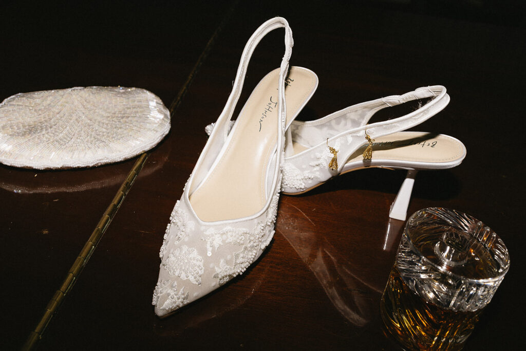 A pair of elegant white lace heels with intricate beading sit on a dark wooden surface next to a beaded clutch and a glass of amber liquid, possibly whiskey.