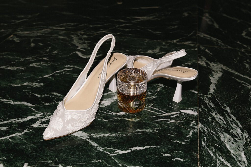 A pair of elegant white slingback heels with floral embroidery sit on dark green marble, with a glass of whiskey placed between them.
