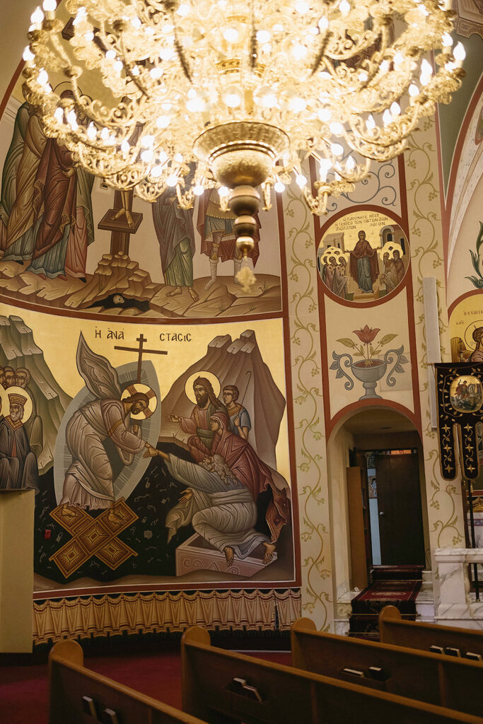 A chandelier hangs above pews in a church adorned with colorful religious murals depicting biblical scenes and figures, with intricate gold accents and arched doorways visible.
