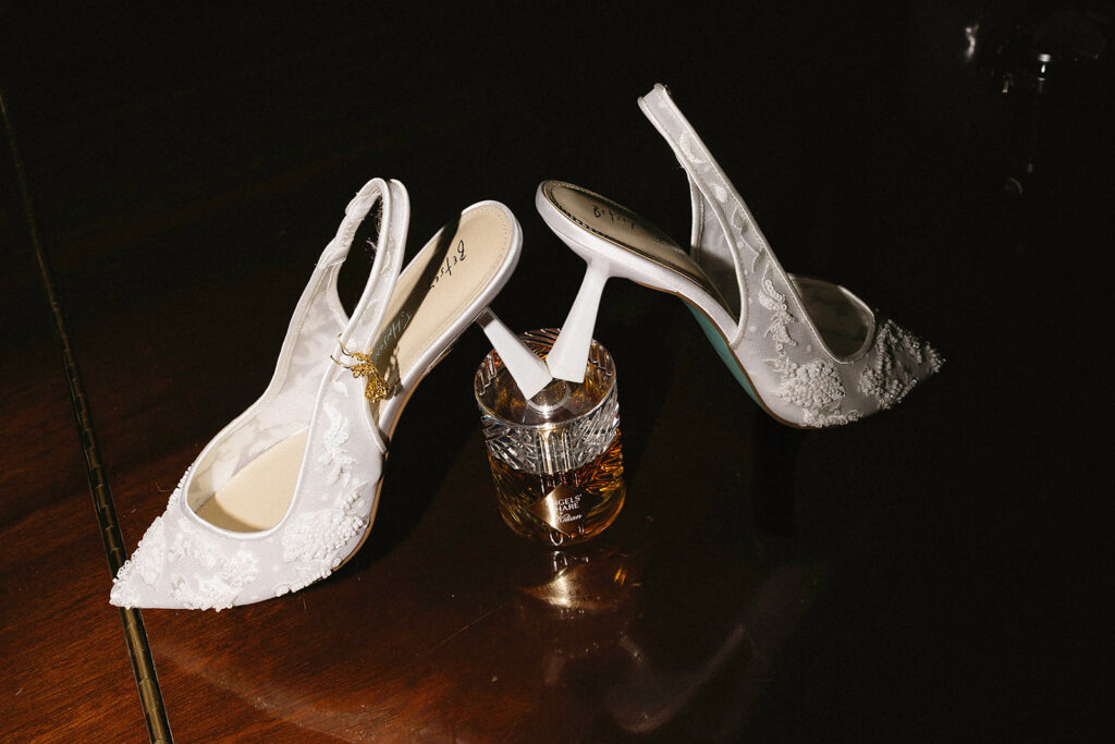 A pair of elegant white lace high-heeled shoes are balanced on the rim of a glass of amber-colored whiskey, placed on a dark wooden surface.