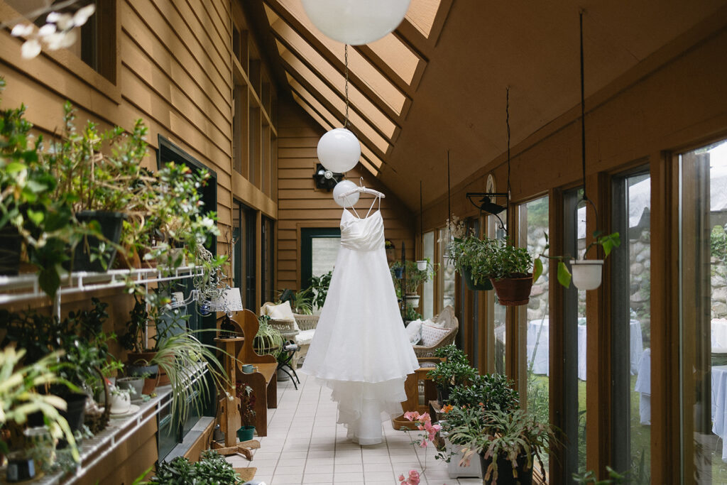 A white wedding dress hangs from the ceiling in a sunlit, plant-filled hallway with large windows, potted plants, and round paper lanterns overhead.