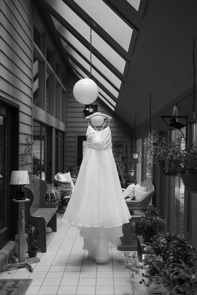 A white wedding dress hangs from a light fixture in a sunlit, plant-filled hallway with windows and skylights. The hallway features benches and cozy seating, creating a peaceful, inviting atmosphere.