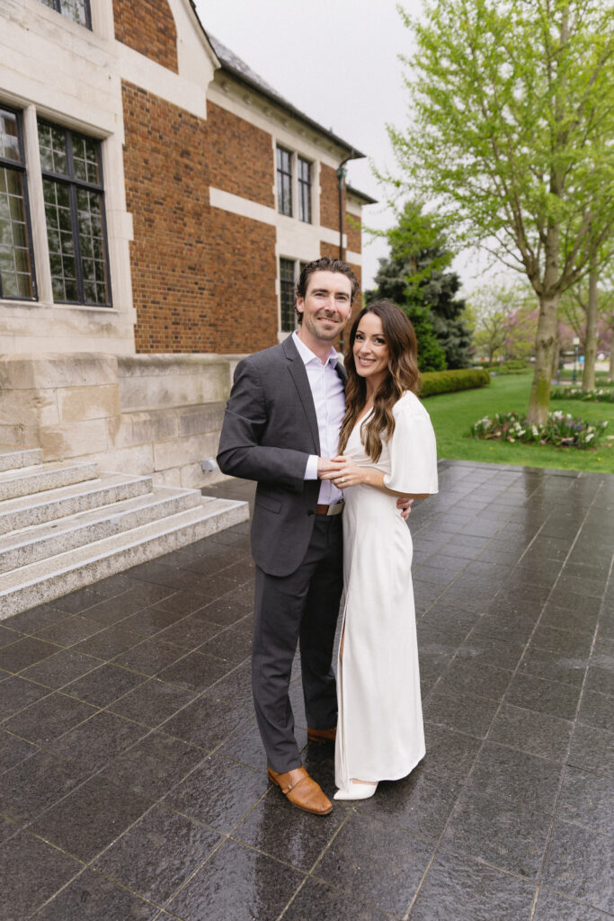 A couple stands close together outside a brick and stone building, smiling at the camera. The man wears a dark suit and brown shoes; the woman wears a white dress. Green trees and a garden are visible in the background.