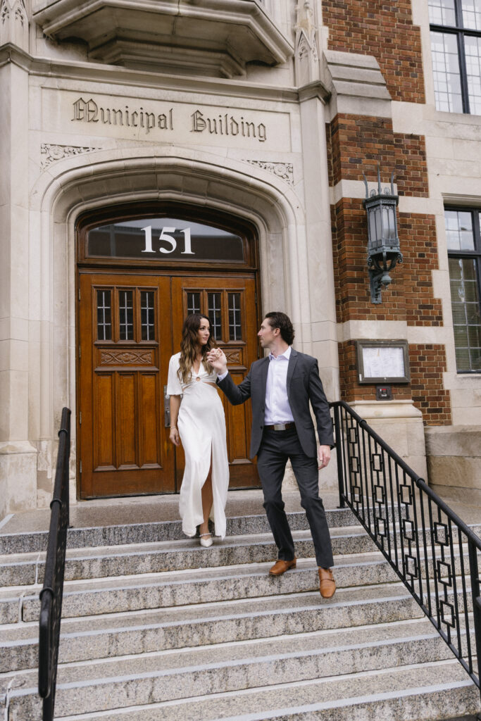 A couple dressed formally holds hands and walks down the steps of a historic building with a sign above the door reading Municipal Building and the number 151.