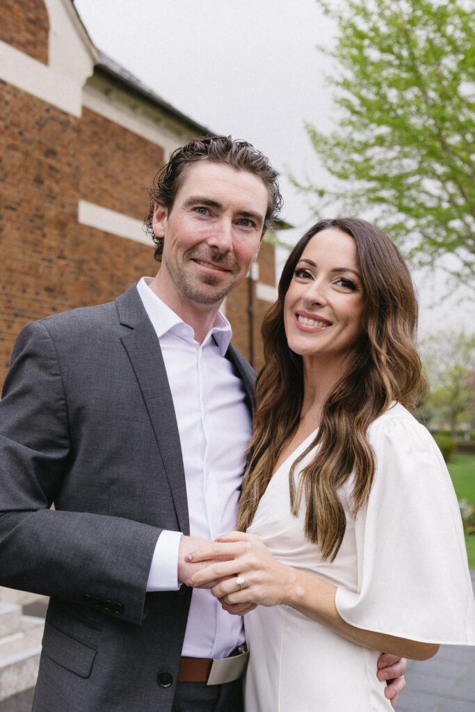A smiling couple in formal attire pose outdoors, the man in a gray suit and white shirt, the woman in a white dress. They hold hands, with greenery and a brick building in the background.