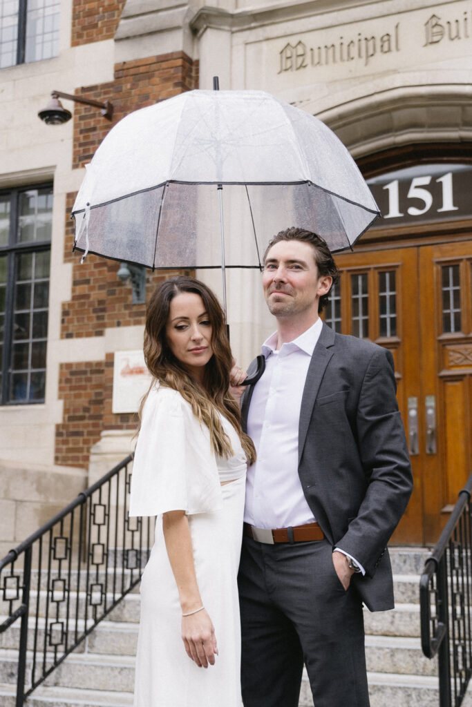 A couple stands on stone steps outside a building, with the man in a dark suit holding a clear umbrella over them. The woman wears a white dress; both look toward the camera. The building has large wooden doors and reads Municipal Building.