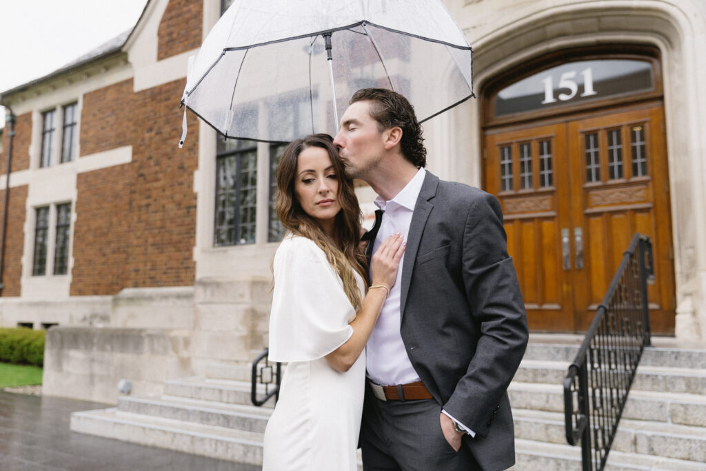 A couple stands close together under a clear umbrella in front of a building with wooden doors and the number 151. The man, in a suit, kisses the womans forehead while she wears a white dress and looks at the camera.
