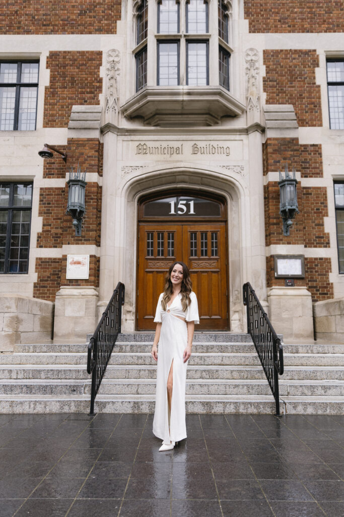 A woman in a white dress stands smiling on the steps outside a brick Municipal Building with wooden doors and the number 151 above the entrance.