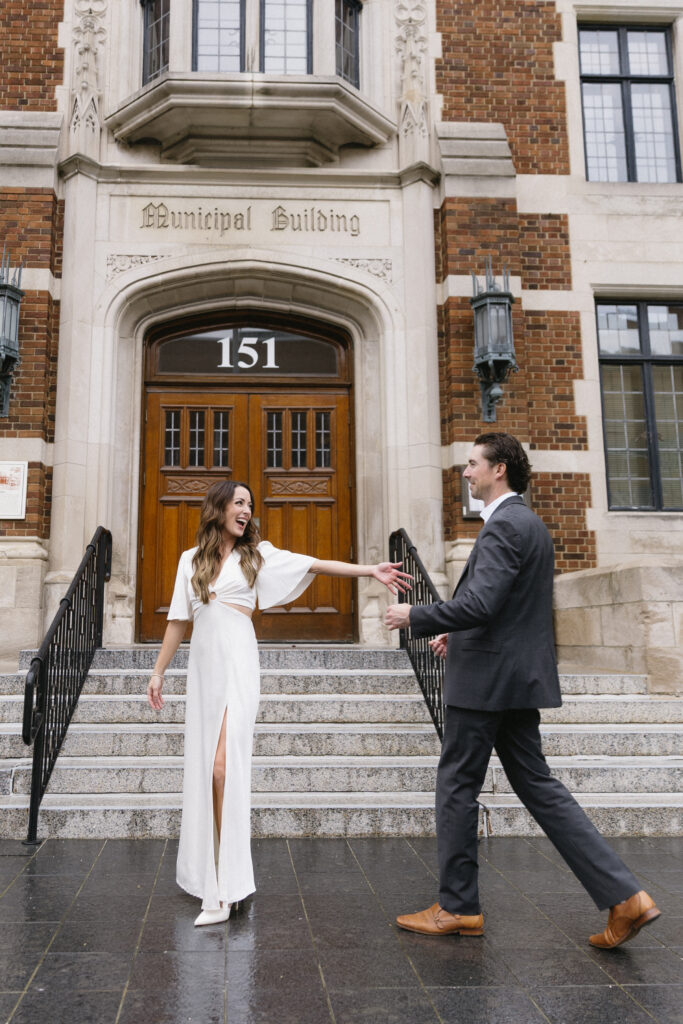 A woman in a white dress and a man in a suit smile and hold hands while standing on the steps of a historic municipal building with the number 151 above the door.