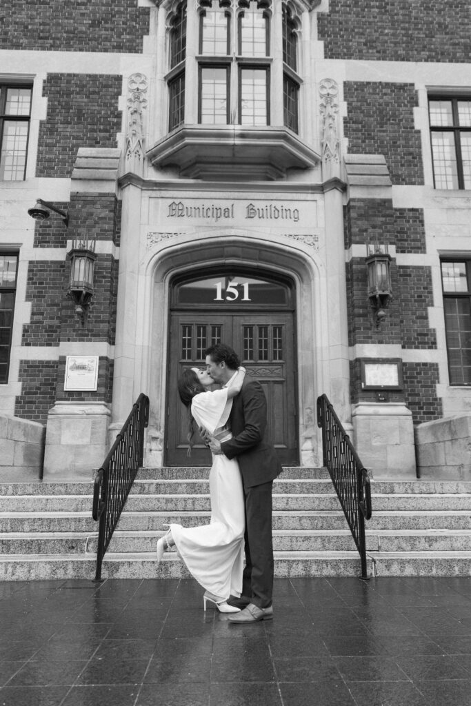 A couple dressed formally shares a joyful kiss in front of a grand building labeled Municipal Building, with the person in white being lifted slightly off the ground on the steps.