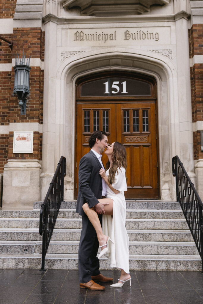 A couple dressed formally stands on the steps of a building labeled Municipal Building and 151. The woman playfully lifts one leg as they smile and gaze at each other affectionately.