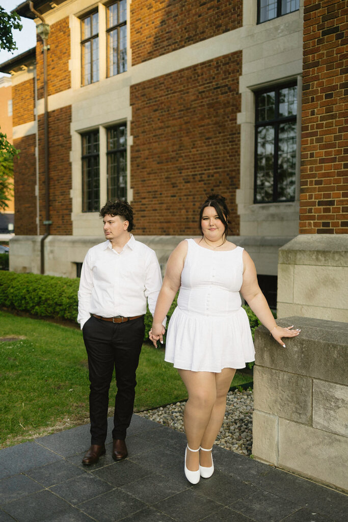 Two people stand outside a brick building. The woman in front wears a white dress and heels, smiling, while the man behind her in a white shirt and black pants looks to the side. They are holding hands.