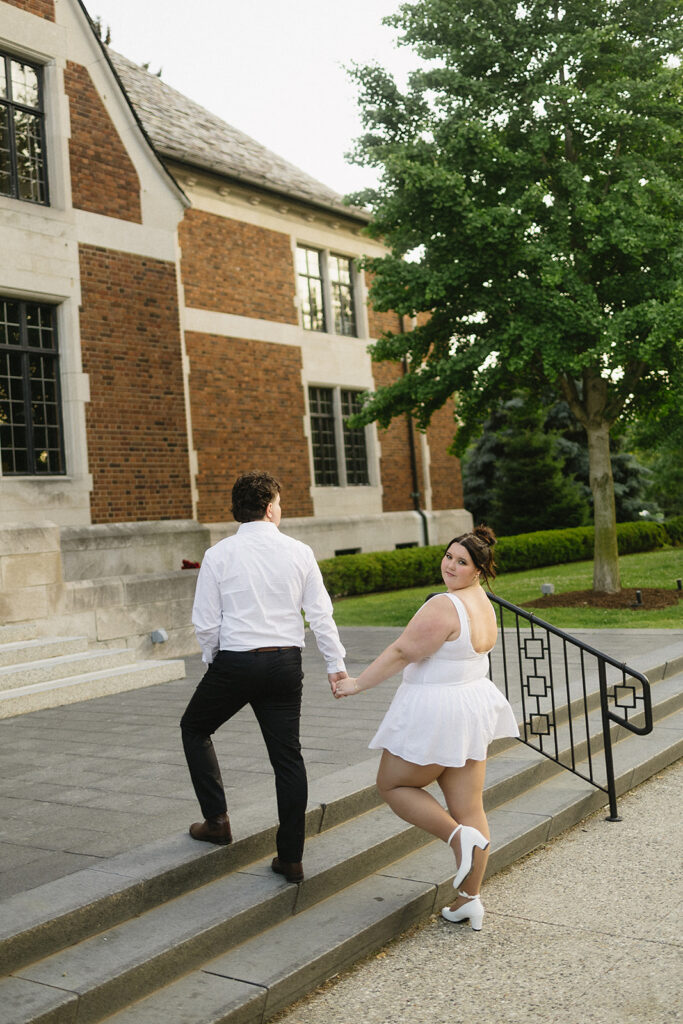 A couple dressed in white holds hands while walking up outdoor steps. The woman looks back and smiles at the camera, while the man faces away. They are near a brick building and green trees.