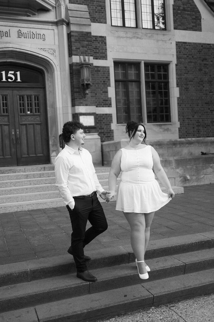 A couple holds hands while standing on steps in front of a brick building. The woman wears a white dress and heels, and the man wears a white shirt and dark pants. Both look at each other and smile. The photo is in black and white.