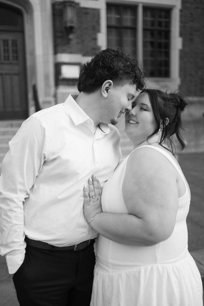A couple stands close together, smiling affectionately as the man leans in to touch his forehead to the woman’s. Both wear white outfits and stand in front of a brick building. The image is in black and white.