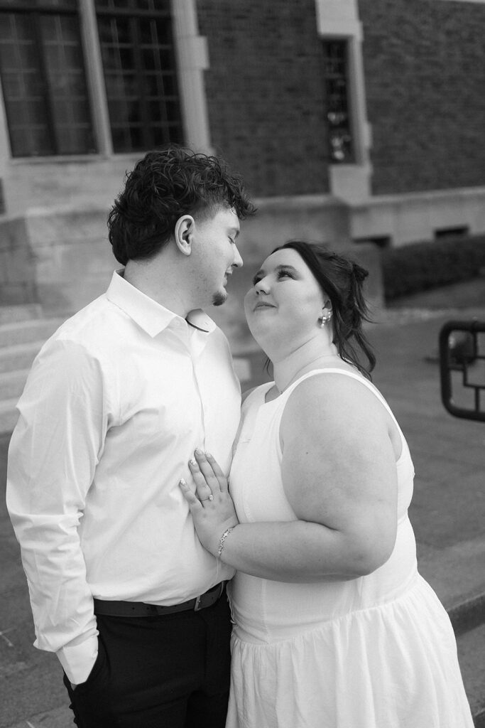 A couple stands closely together outside a brick building, gazing into each others eyes with affectionate expressions. The woman has her hand on the mans chest, and both are dressed in light-colored clothing.