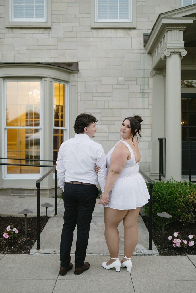 A couple stands hand in hand outside a stone building, both dressed in white. The woman looks over her shoulder and smiles at the camera, while the man faces her, with a warm light shining through a window behind them.