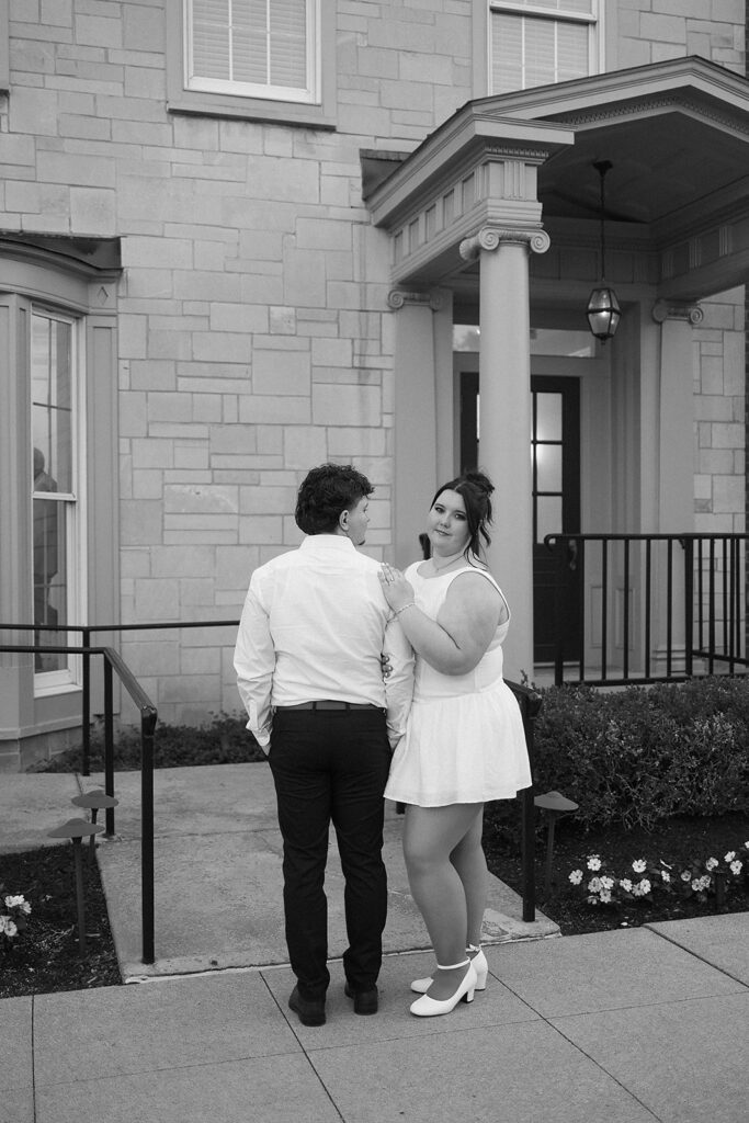 A couple stands outside a stone building. The woman, in a white dress and heels, smiles at the camera with her hand on the mans shoulder. The man, in dark pants and a white shirt, faces away from the camera.