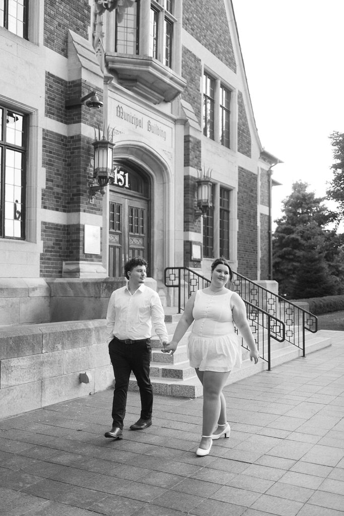 A couple dressed in white holds hands and walks outside a brick municipal building. They smile at each other, and the scene has a soft, candid feel. The building’s address, 151, is visible above the entrance.