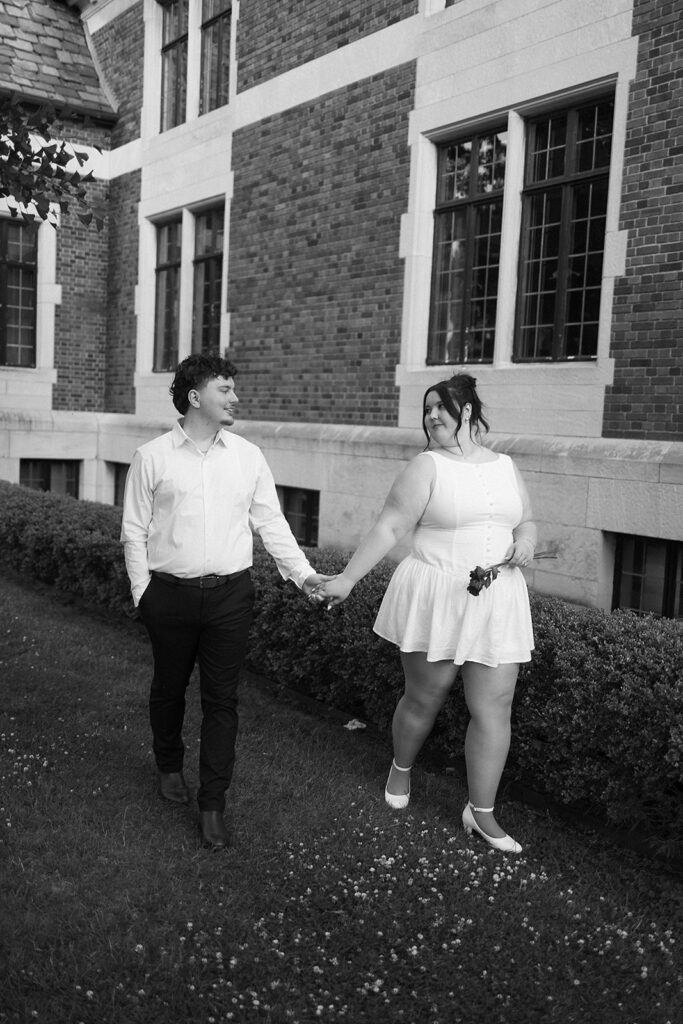 A couple holding hands and smiling at each other as they walk outside along a trimmed hedge in front of a brick building with large windows. The image is in black and white.