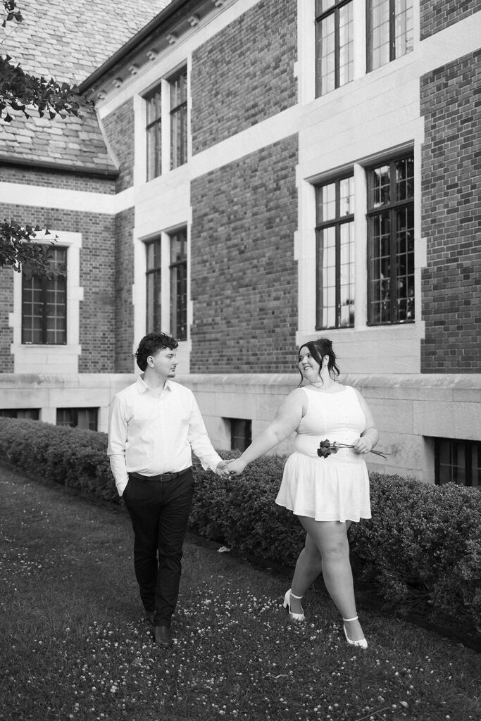 A couple holds hands and smiles at each other while walking outside a brick building with large windows. The woman wears a white dress and heels, and the man wears dark pants and a white shirt. The photo is in black and white.
