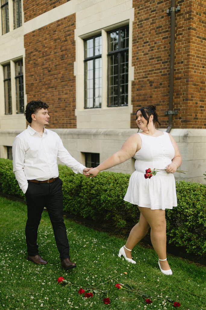 A couple holding hands walks on grass outside a brick building. The woman, in a white dress and heels, smiles at the man and holds roses. The man, in a white shirt and black pants, looks back at her. Roses are scattered on the ground.