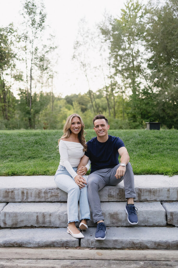 A smiling couple sits on stone steps outdoors. The woman, wearing a white off-shoulder top and light jeans, holds hands with the man, who is in a navy t-shirt and gray pants. Green grass and trees are in the background.